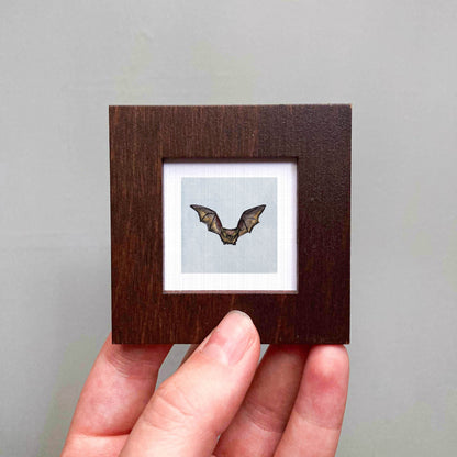 A person's hand is holding a small wooden frame containing a picture of a bat.