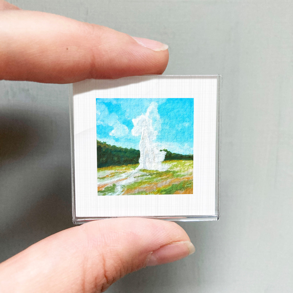 A person's hand holding a small square picture of a landscape with a white geyser in the background.