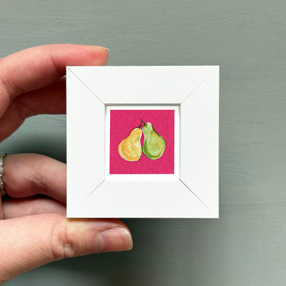 A hand is holding a small white square frame containing a painting of two pears.
