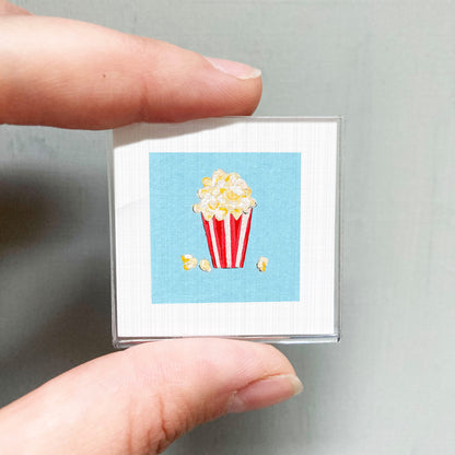 A hand is holding a small square with a picture of a red and white striped popcorn container on a blue background.