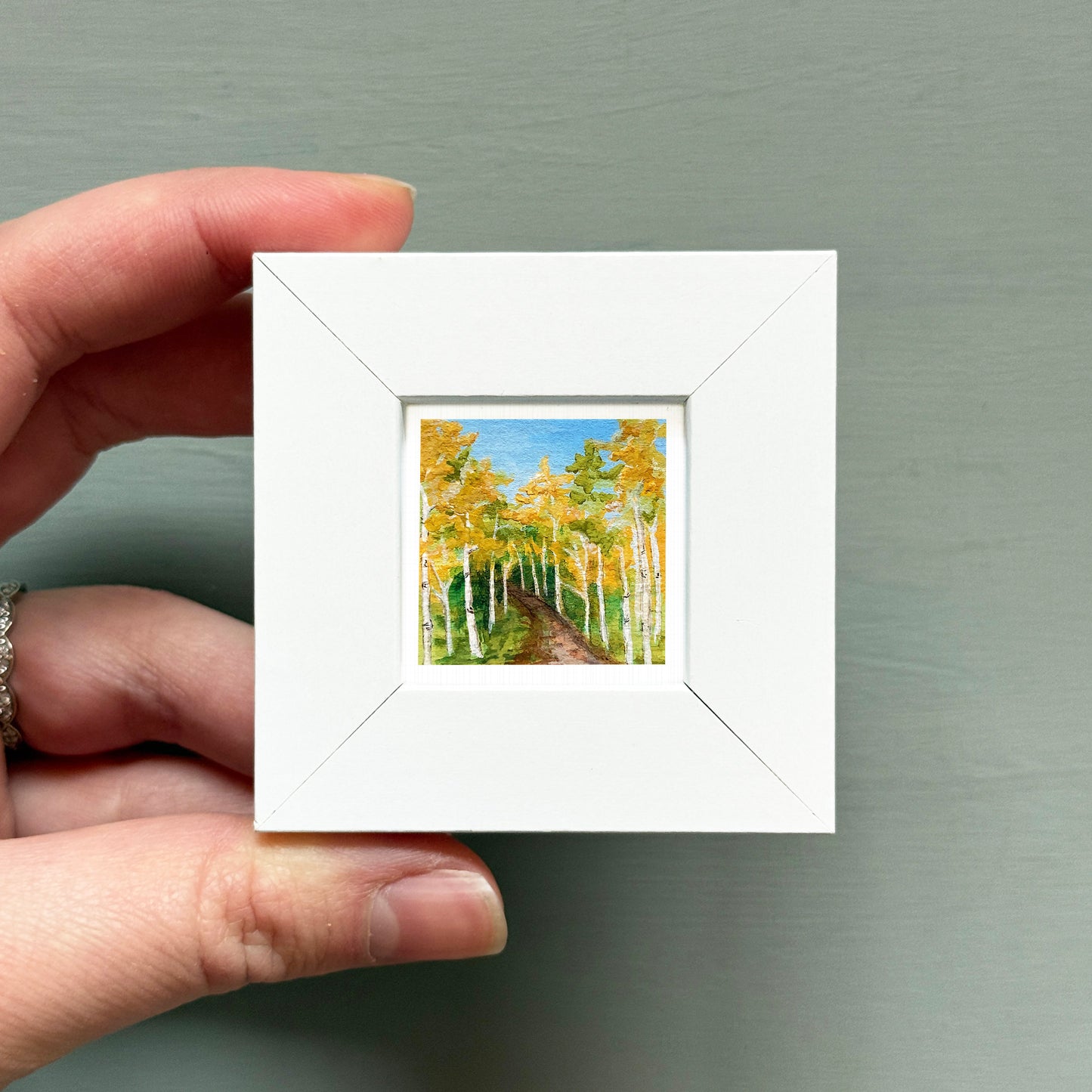 A hand is holding a small white square frame containing a painting of a forest with yellow trees.