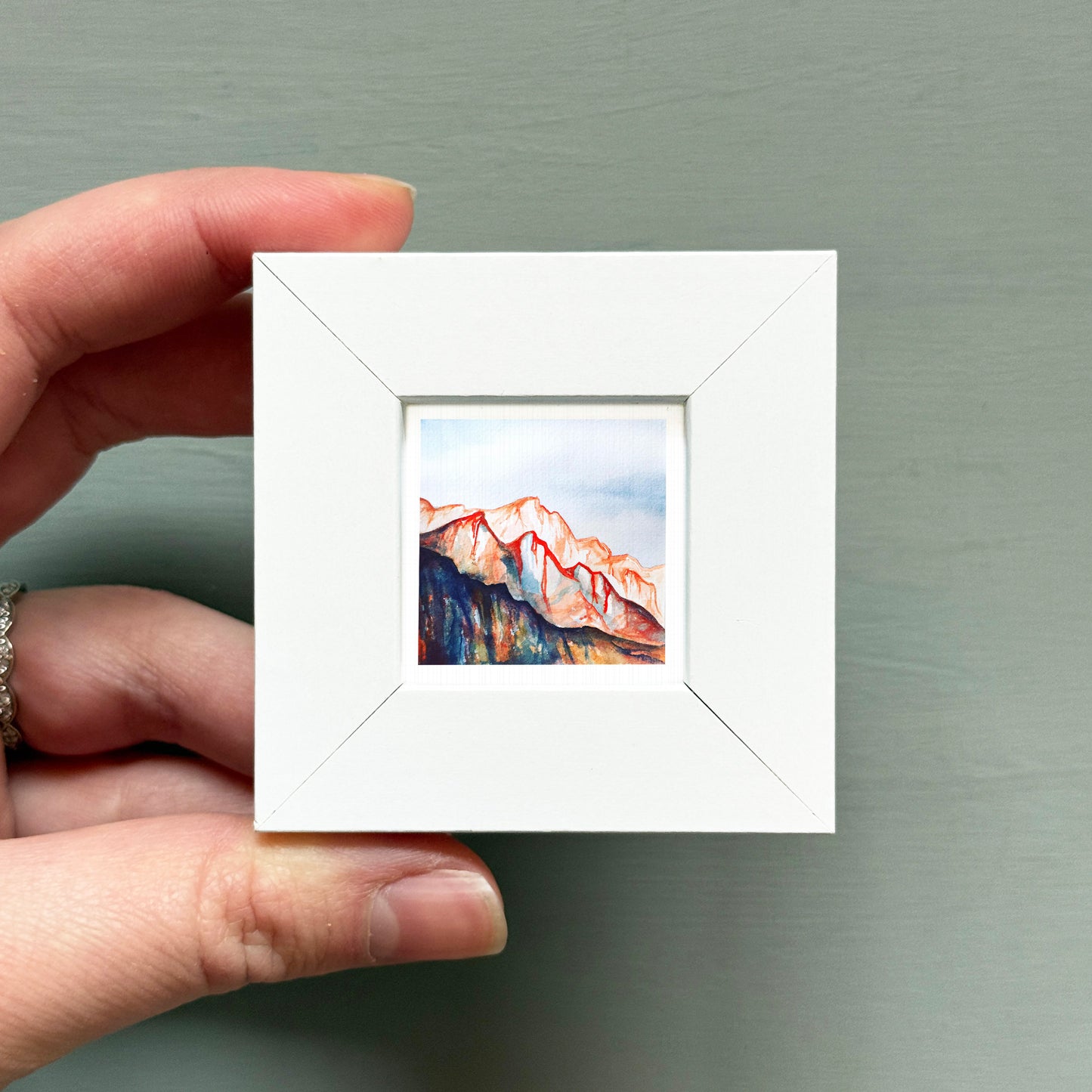 A hand is holding a small white square frame containing a watercolor painting of a mountain landscape.