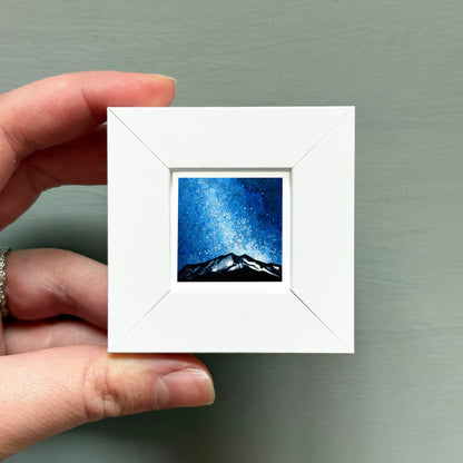 A hand is holding a small white square frame containing a painting of a mountain landscape under a starry night sky.