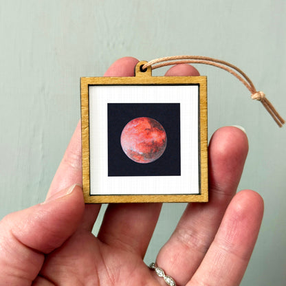 A person's hand holding a small wooden frame containing a photograph of a red planet, likely Mars, against a black background.