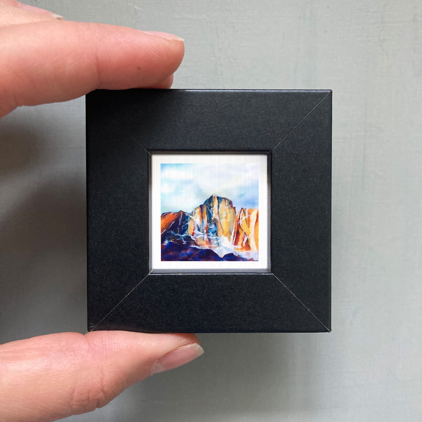 A person's hand is holding a small black frame containing a framed painting of a mountain landscape.
