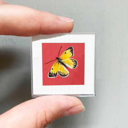 A hand is holding a small square card with a vibrant painting of a yellow butterfly on a red background.