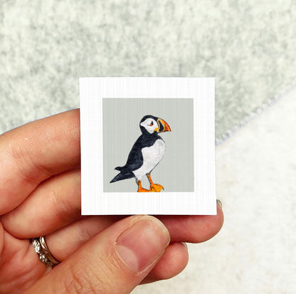 A person's hand holding a small square picture of a puffin bird.