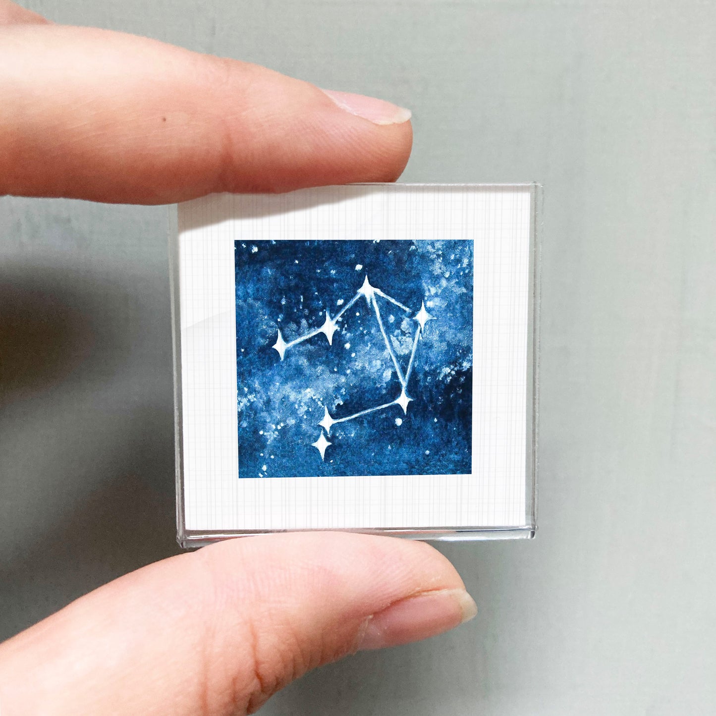 A person's hand holding a small square-shaped object with a blue and white starry night sky design on it.