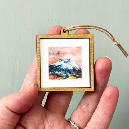 A hand holding a small wooden frame containing a watercolor painting of a mountain landscape.