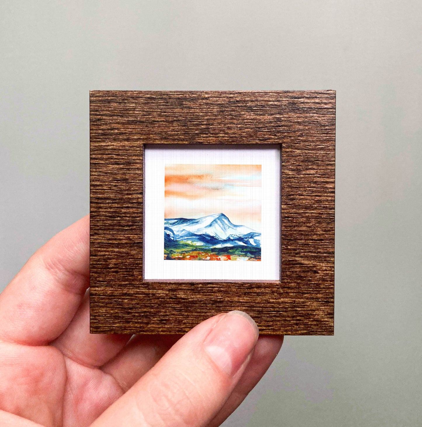 A hand is holding a small wooden frame containing a framed landscape painting depicting a mountain scene.