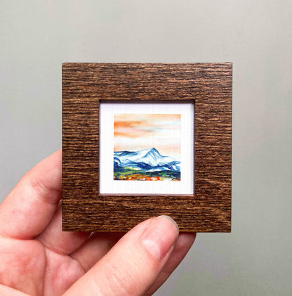 A hand is holding a small wooden frame containing a framed landscape painting depicting a mountain scene.