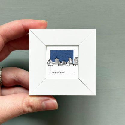 A hand is holding a small white picture frame containing an illustration of a city skyline.