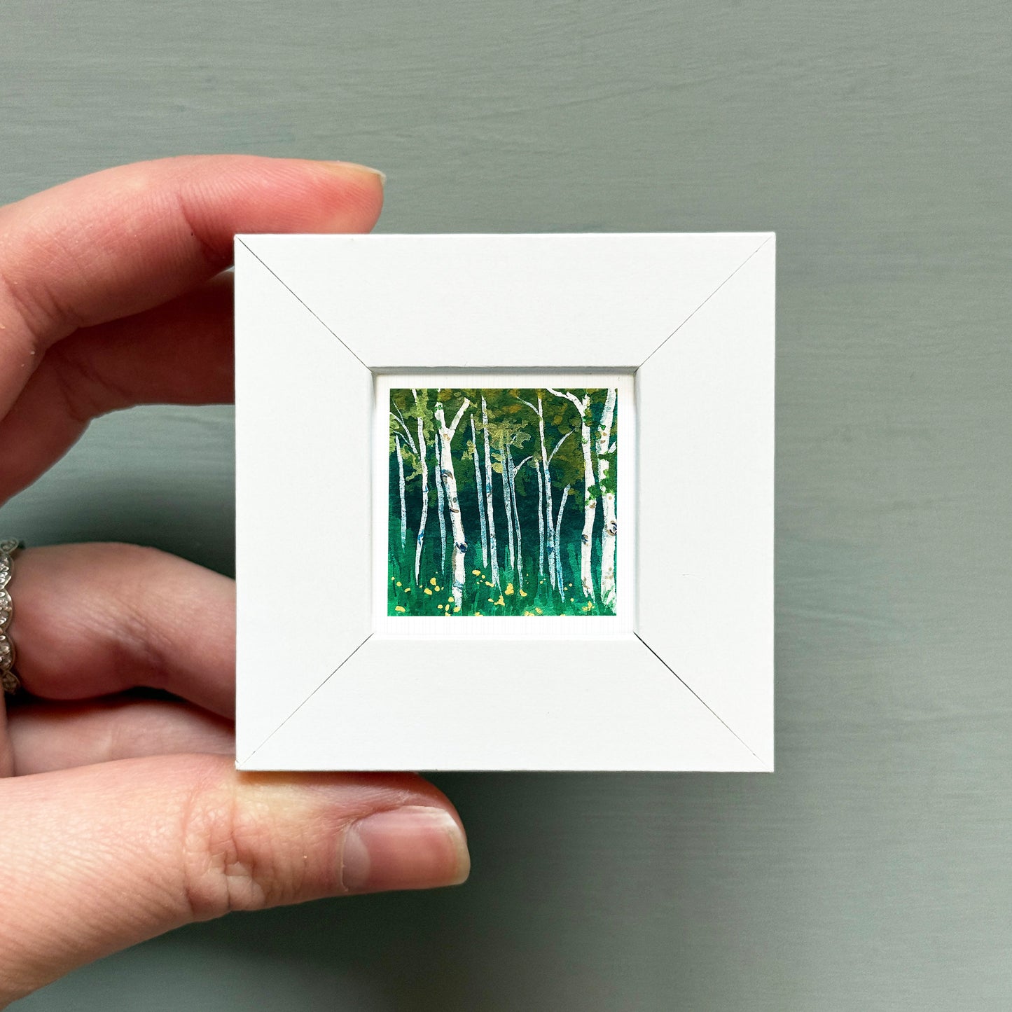 A hand is holding a small white square frame containing a painting of a forest with tall trees and green foliage.