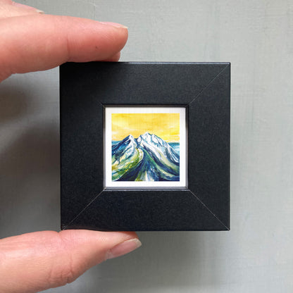 A person's hand holding a small black frame containing a framed painting of a mountain landscape with a yellow sky.