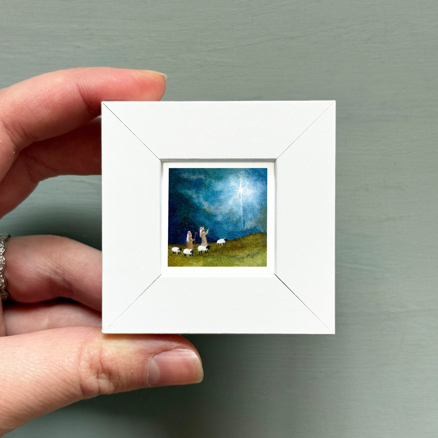 A hand is holding a small white square frame containing a painting of a pastoral scene with sheep grazing on a hill.