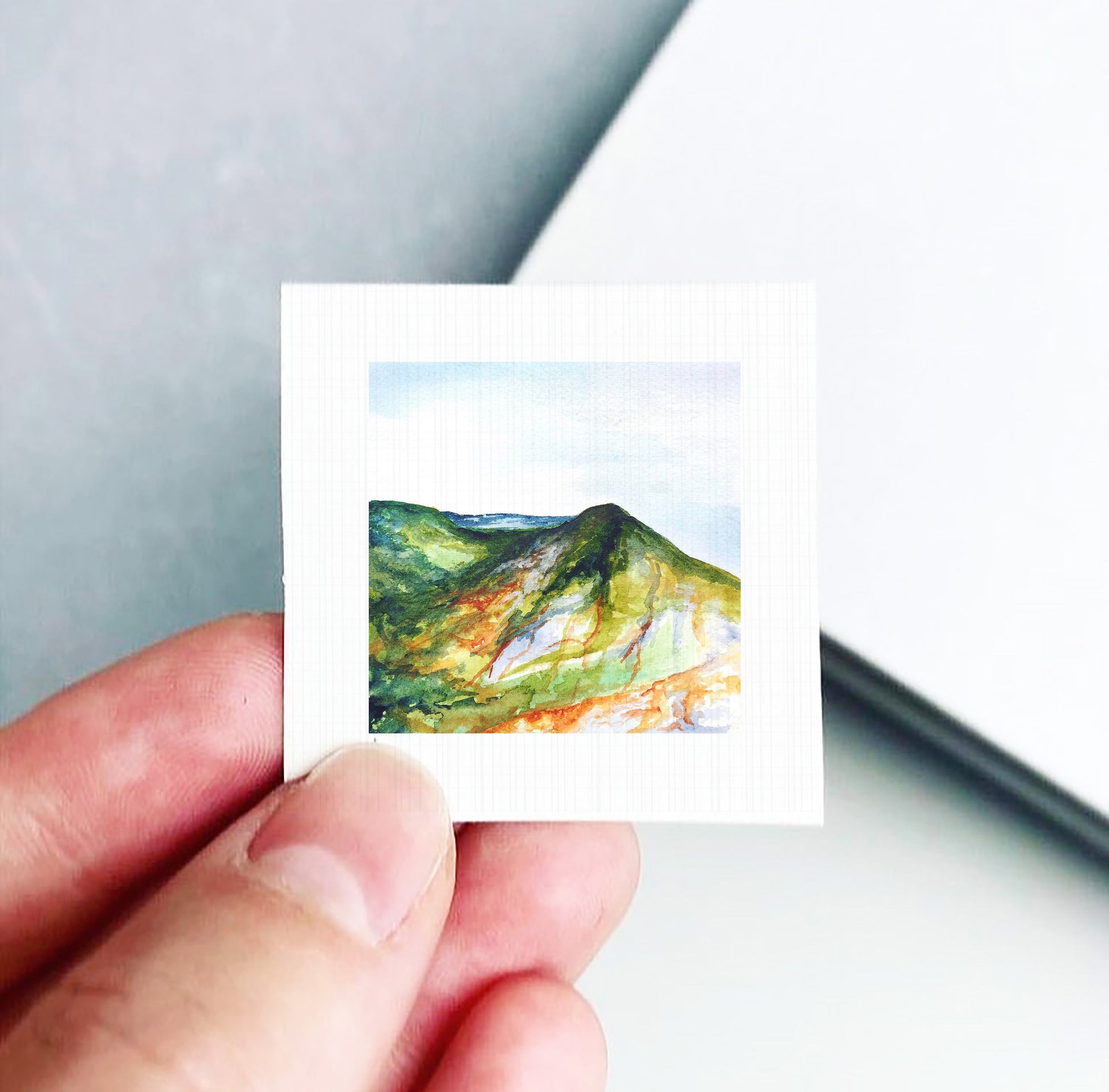 A hand holding a small square artwork depicting a mountain landscape with a green and orange color palette.