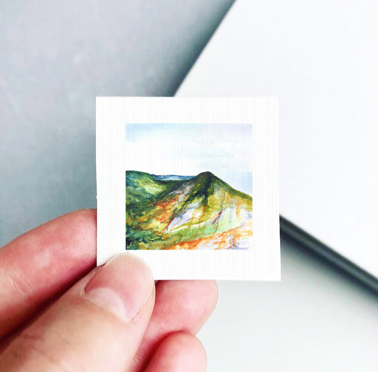 A hand holding a small square artwork depicting a mountain landscape with a green and orange color palette.