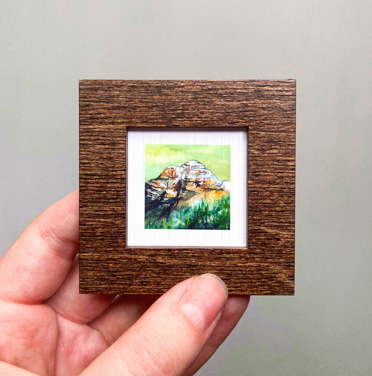 A hand holding a small wooden frame containing a watercolor painting of a mountain landscape.