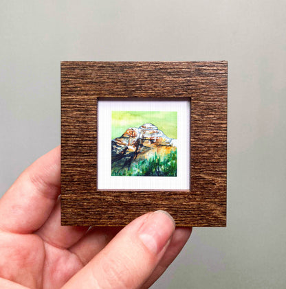 A hand holding a small wooden frame containing a watercolor painting of a mountain landscape.