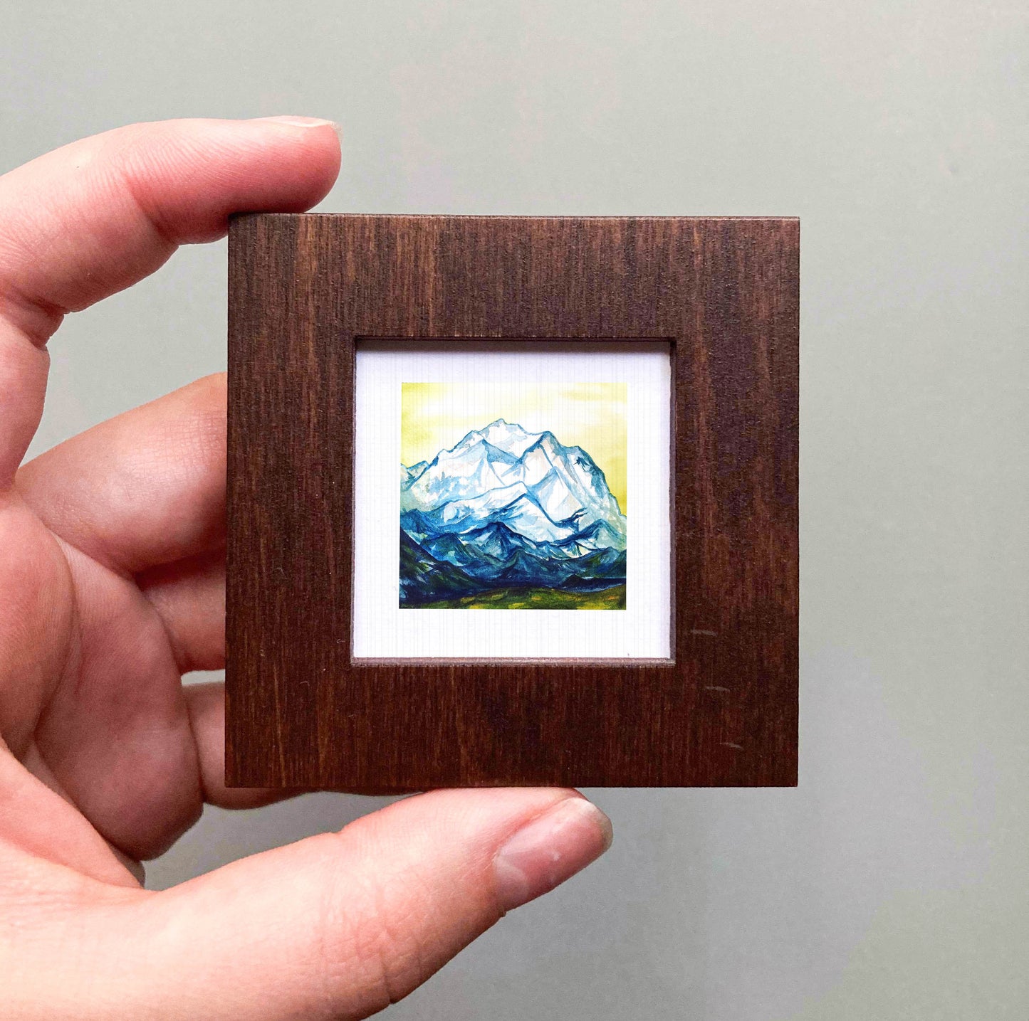 A hand is holding a small wooden frame containing a painting of a mountain landscape.
