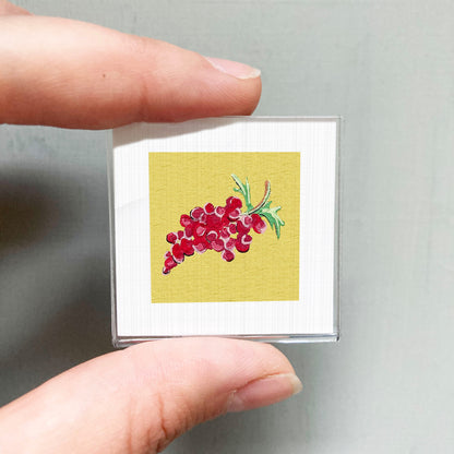 A person's hand holding a small square with a vibrant illustration of red grapes on a yellow background.