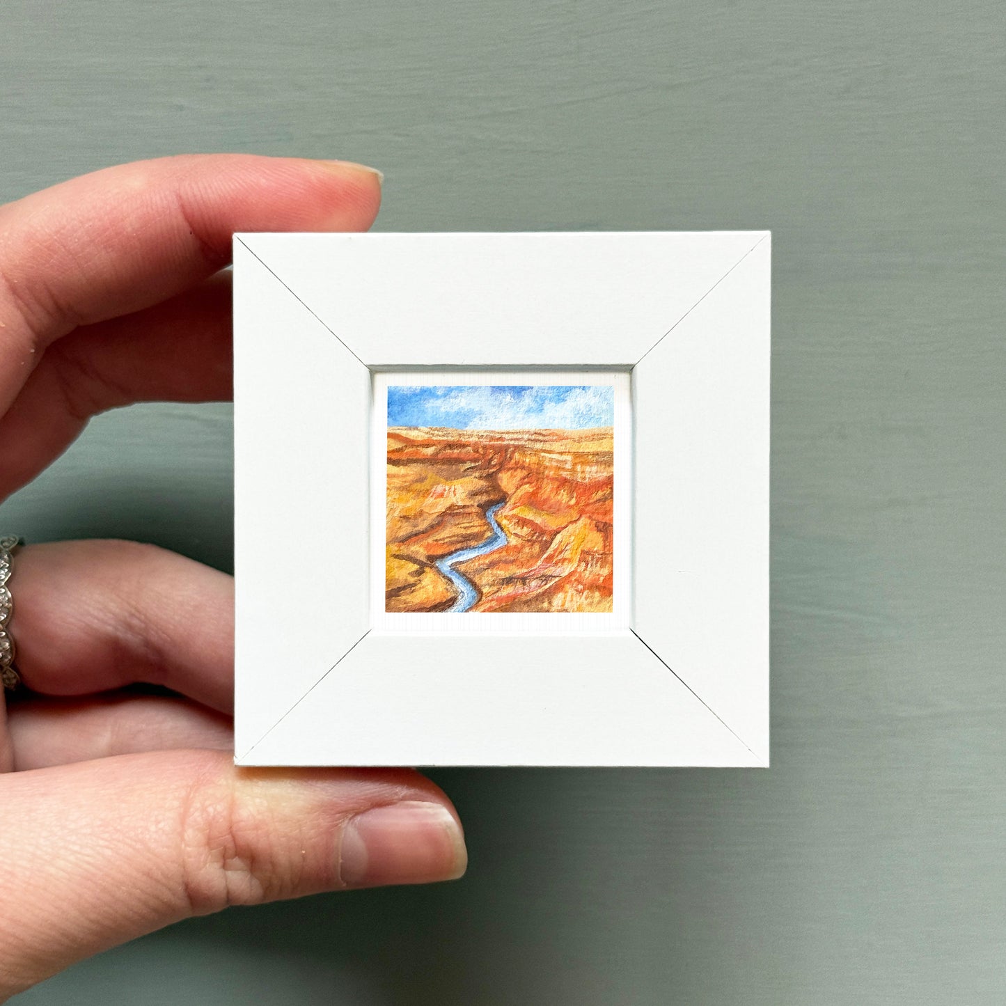 A hand is holding a small white square frame containing a painting of a desert landscape with a winding river.