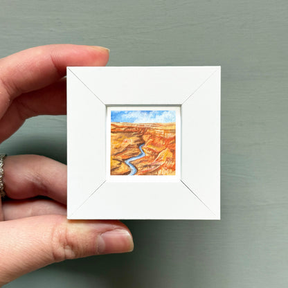 A hand is holding a small white square frame containing a painting of a desert landscape with a winding river.