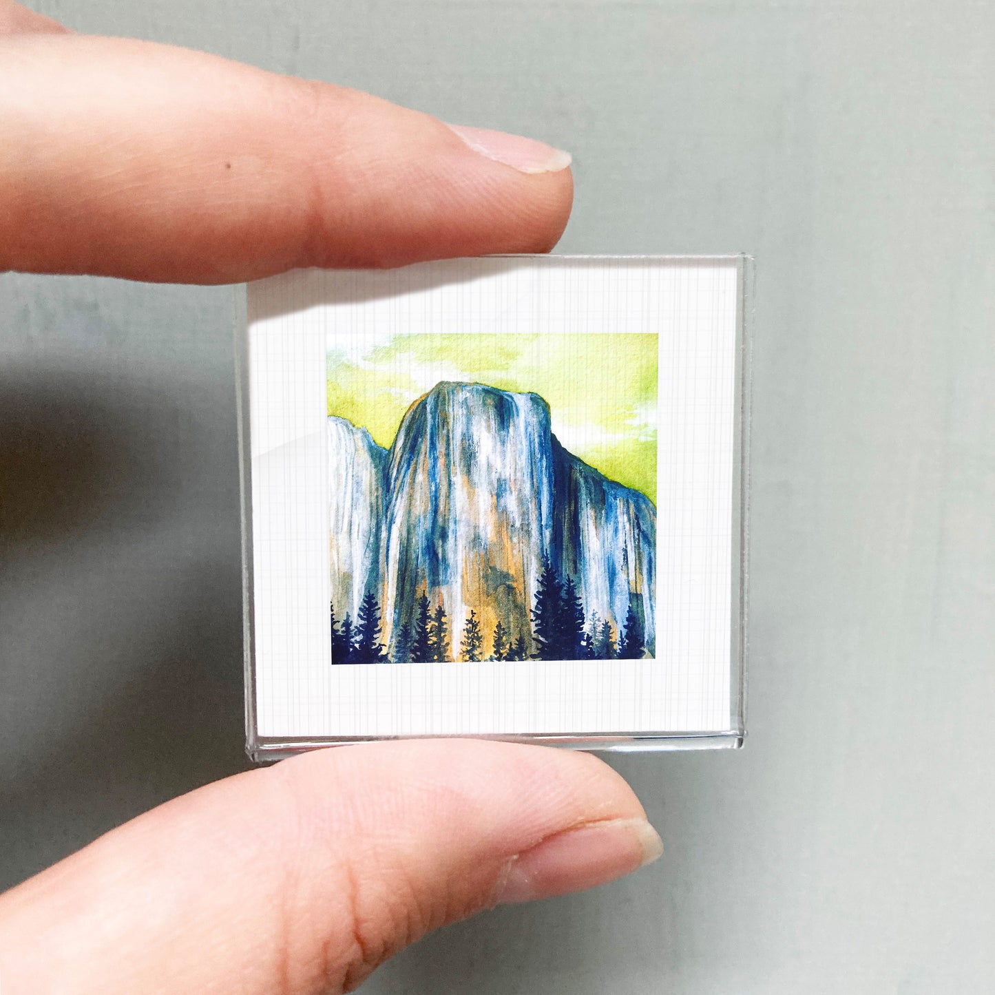 A person's hand holding a small square picture of a mountain landscape with trees and a waterfall.