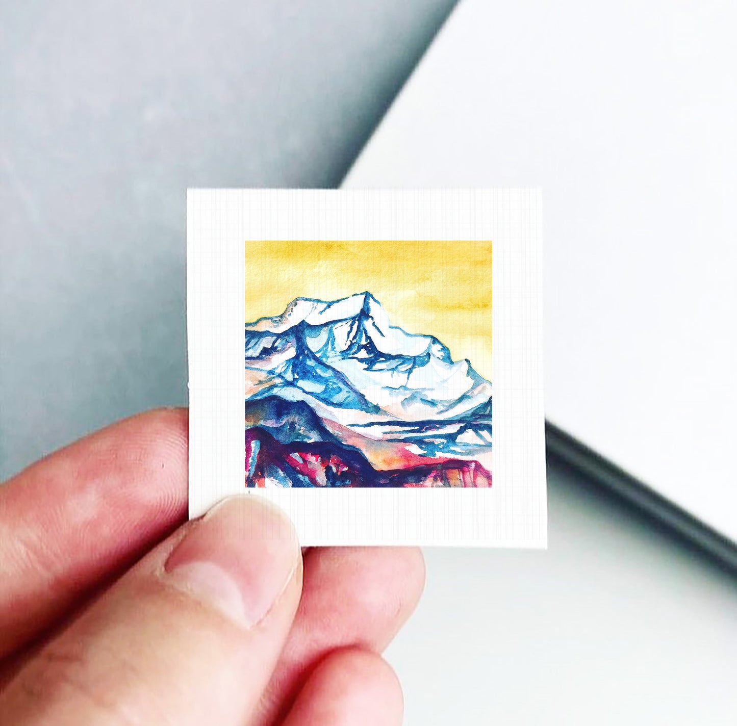 A hand is holding a small square piece of paper with a watercolor painting of a mountain landscape on it.