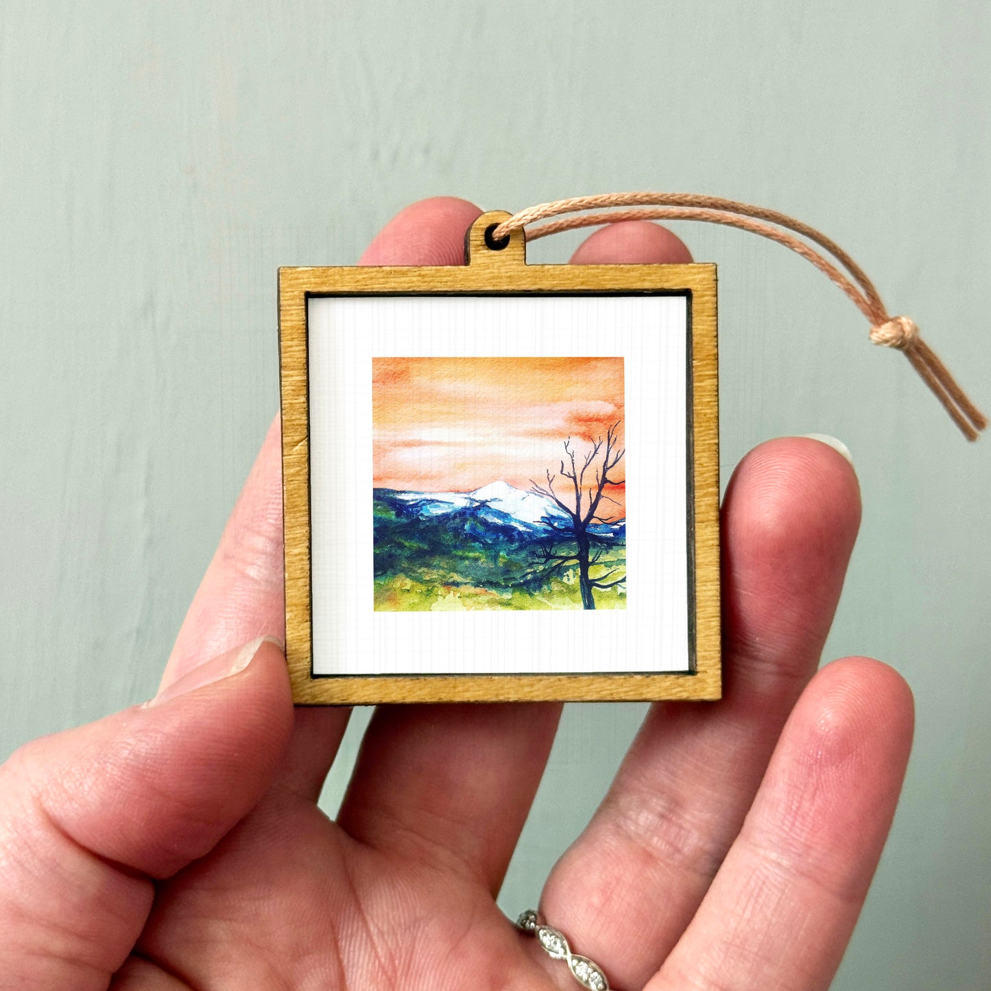 A hand holding a small wooden frame containing a painting of a mountain landscape.