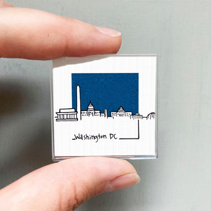A person's hand holding a small square magnet with a drawing of the Washington, D.C. skyline.