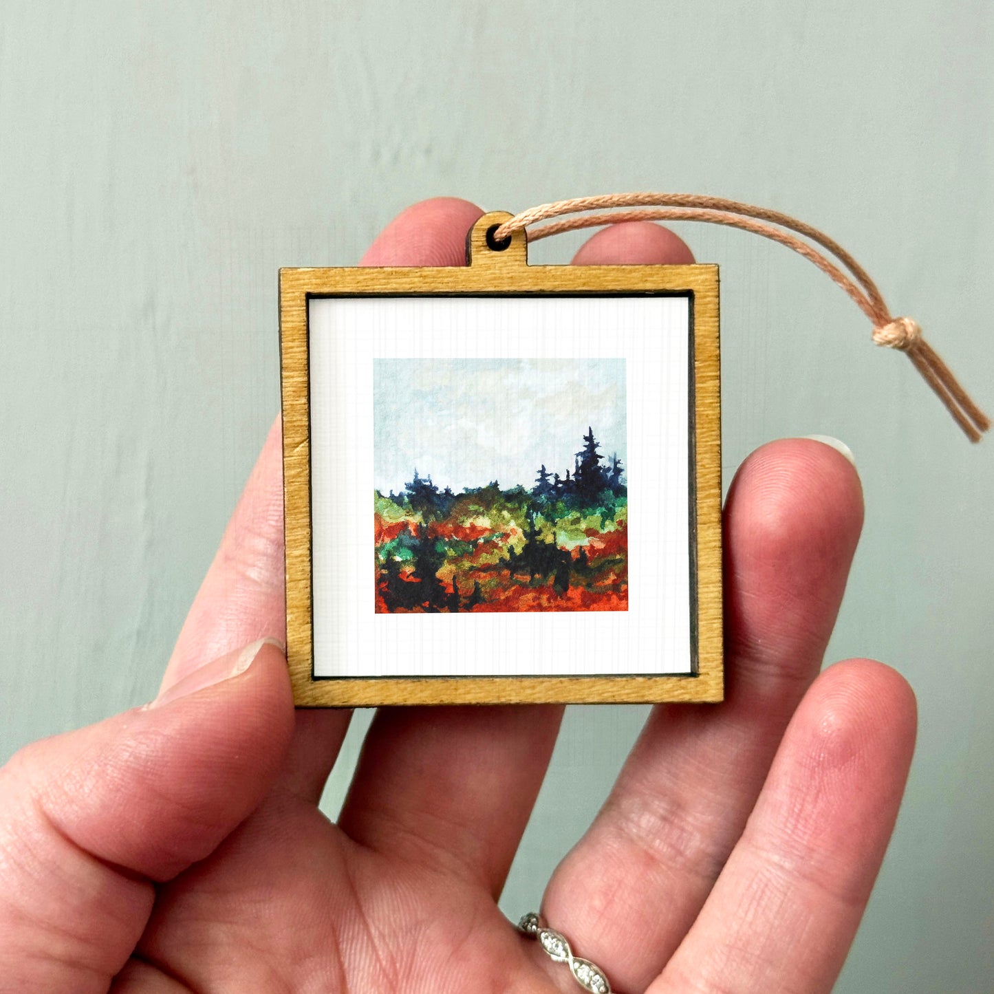 A hand holding a small wooden frame containing a painting of a landscape with trees and a blue sky.
