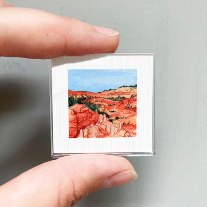 A person's hand holding a small square picture of a desert landscape with red rock formations and a blue sky.