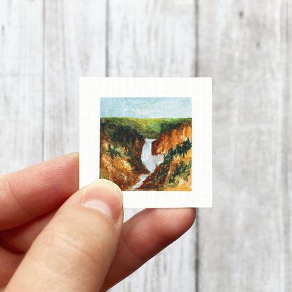 A hand is holding a small square-shaped painting depicting a scenic landscape with a waterfall.