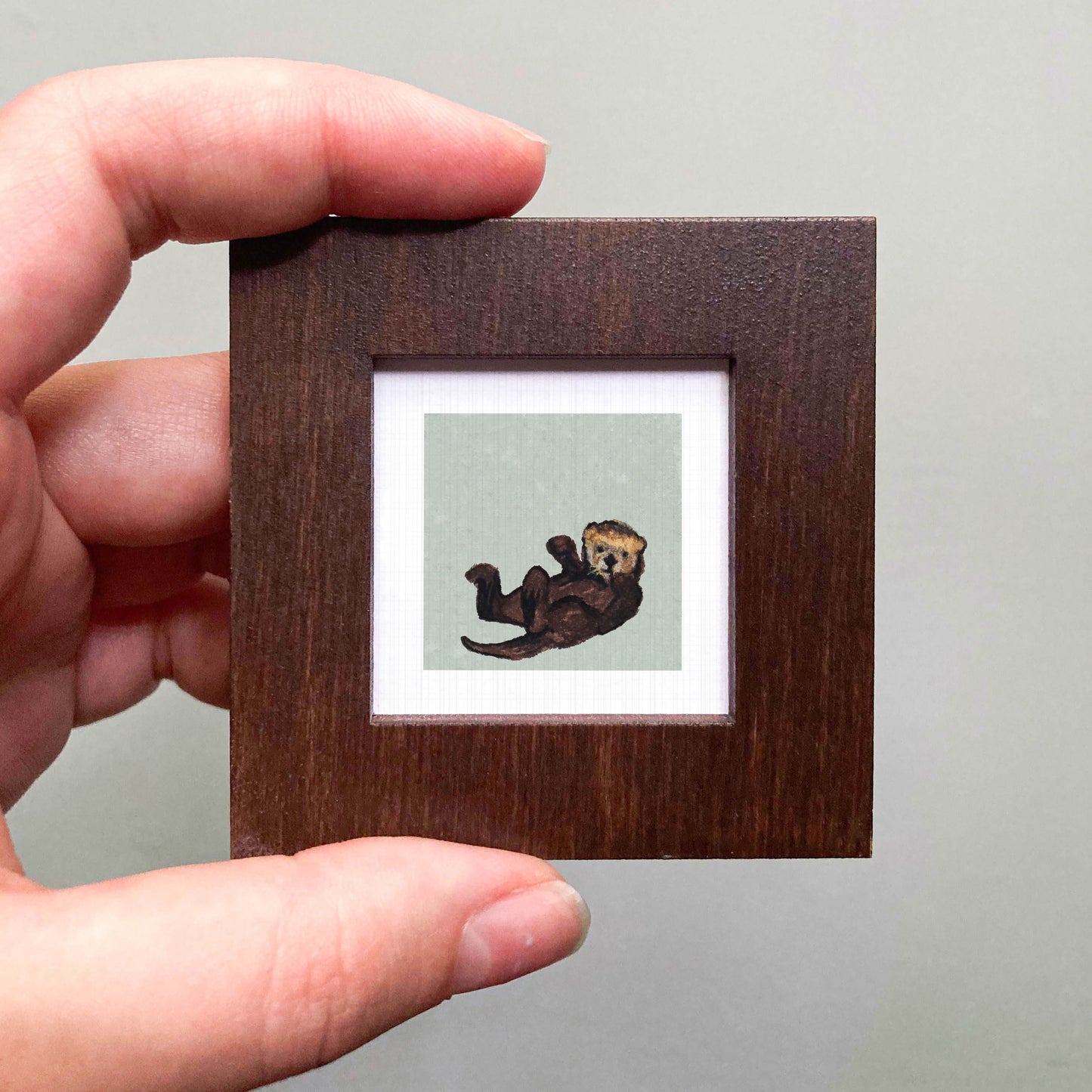A person's hand is holding a small wooden frame containing a picture of an otter.