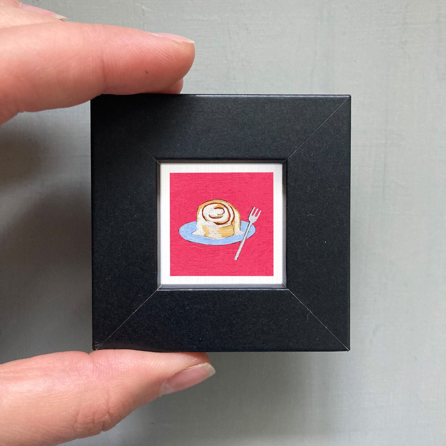 A hand is holding a small black picture frame containing a framed illustration of a cinnamon roll on a plate.