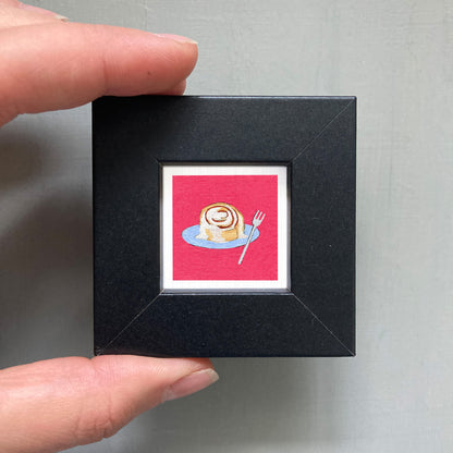 A hand is holding a small black picture frame containing a framed illustration of a cinnamon roll on a plate.