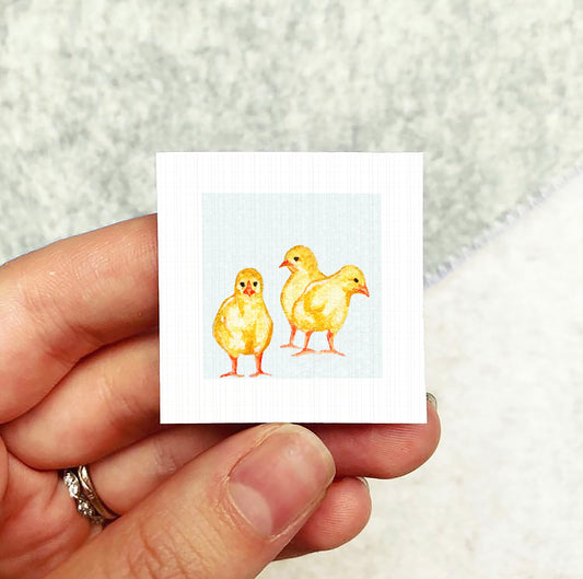 A hand holding a small square card with an illustration of three yellow chicks on it.