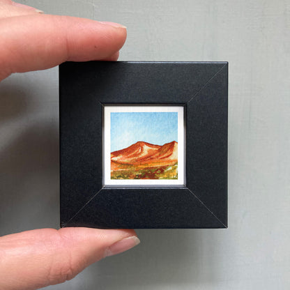A person's hand holding a small black picture frame containing a watercolor painting of a mountain landscape.