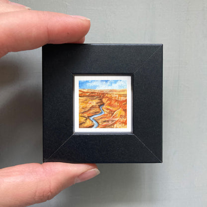A person's hand holding a small black frame containing a miniature painting of a desert landscape with a winding river.