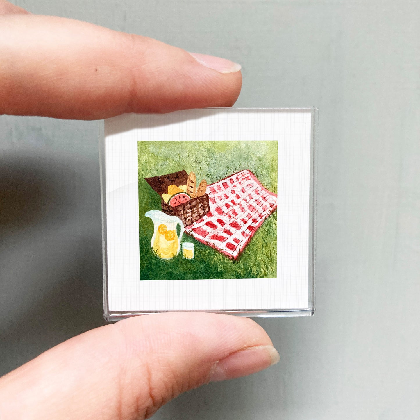 A person's hand holding a small square picture depicting a picnic scene with a basket, a pitcher of lemonade, and a red and white checkered blanket on the grass.