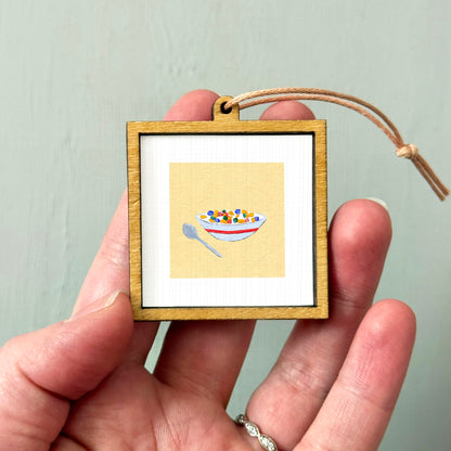 A person's hand holding a small wooden frame containing a picture of a bowl of cereal.