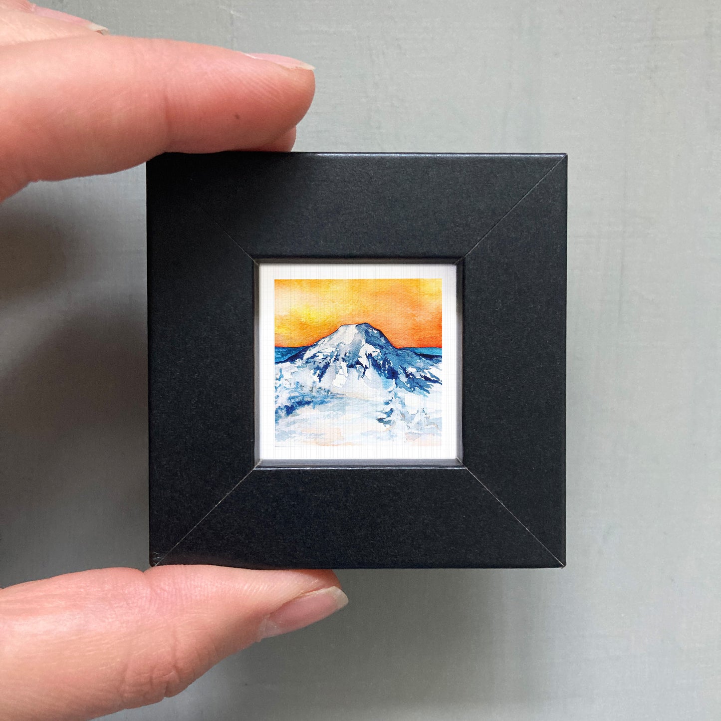 A person's hand holding a small black frame containing a framed painting of a mountain landscape with a vibrant orange and blue sky.