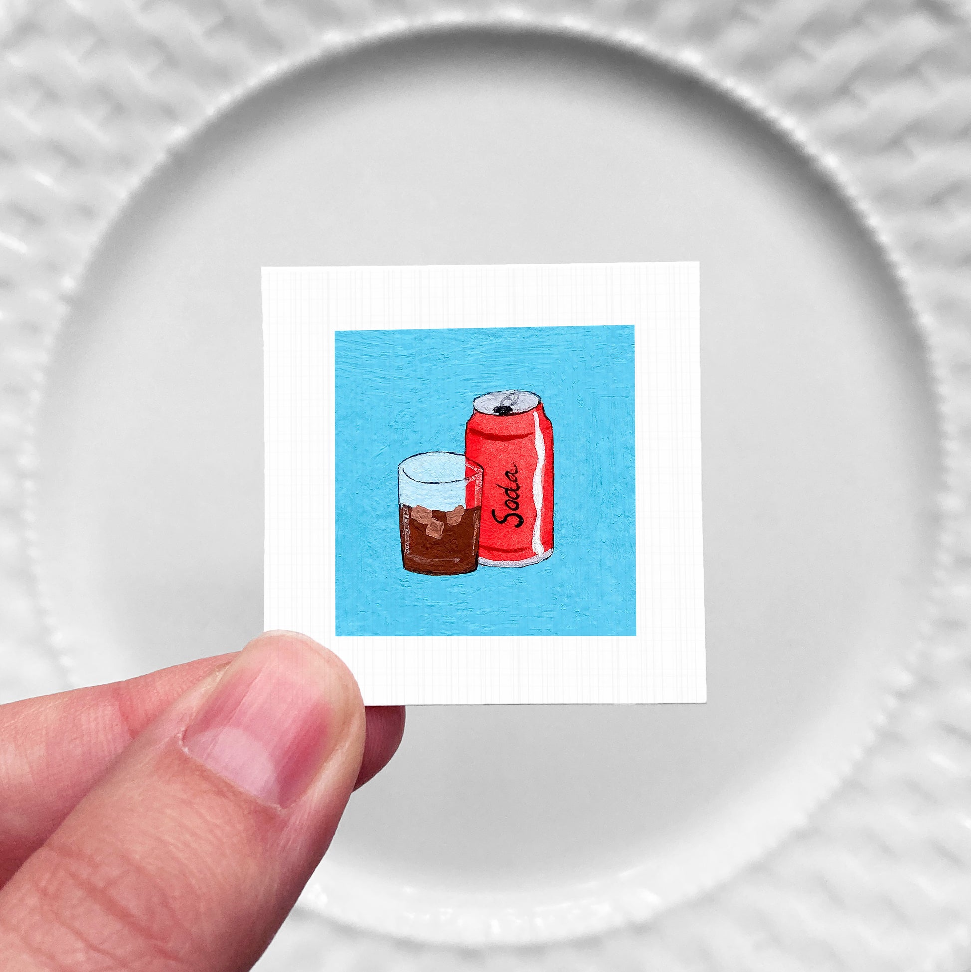 A hand is holding a small square with an illustration of a glass of soda and a can of soda on it.