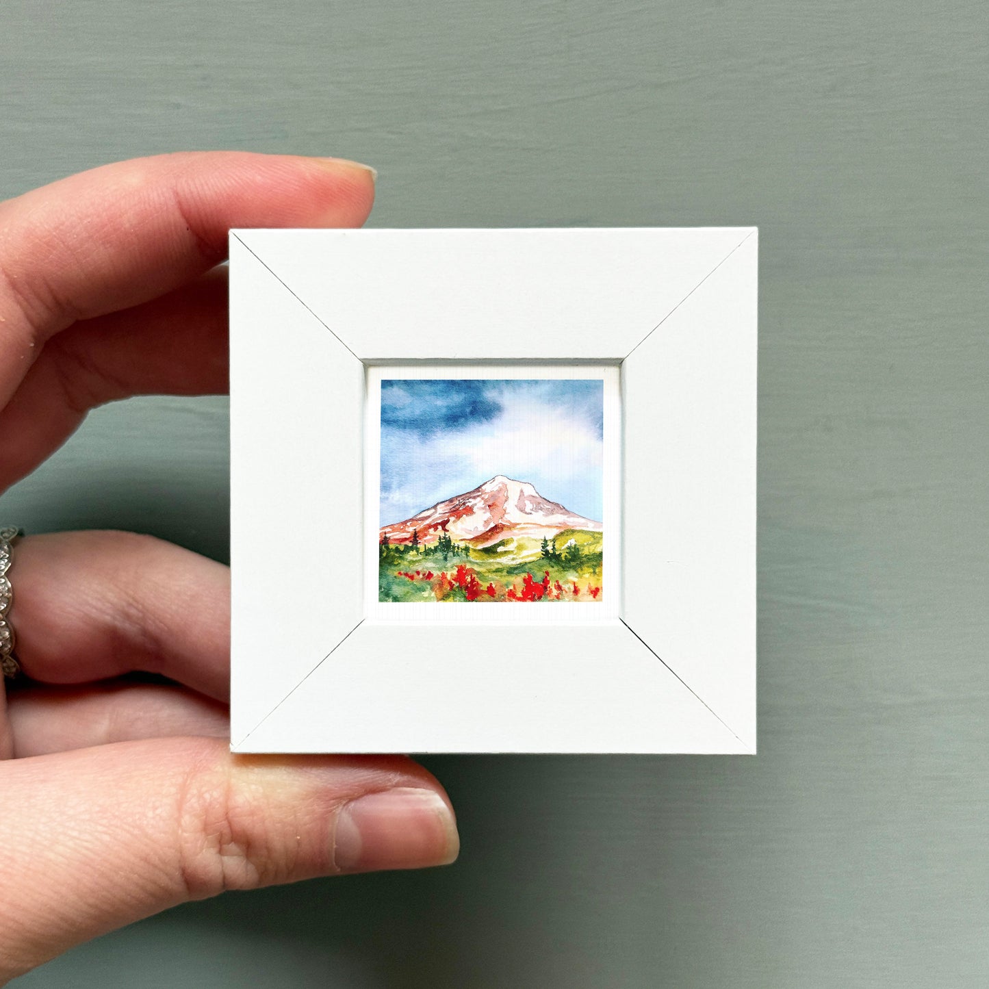 A hand is holding a small white square frame containing a watercolor painting of a mountain landscape.