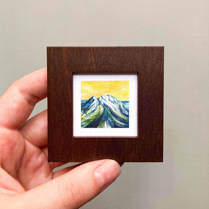 A hand is holding a small wooden frame containing a painting of a mountain landscape with a yellow sky.