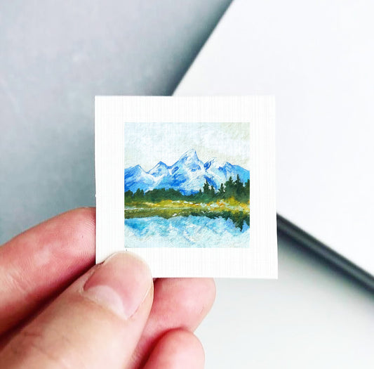 A hand is holding a small square painting depicting a mountain landscape with a lake and trees.