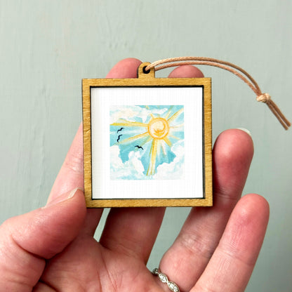 A hand holding a small wooden frame containing a watercolor painting of a sun and clouds.