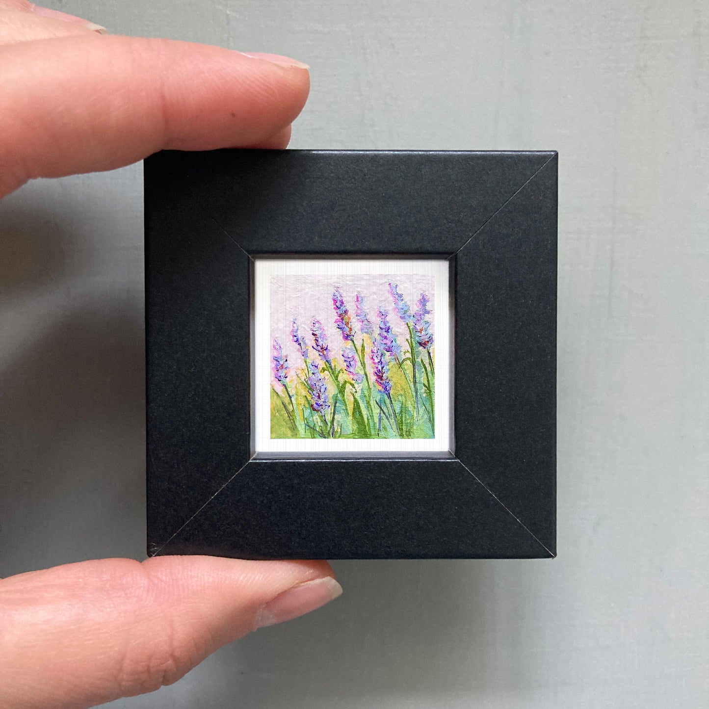 A hand is holding a small, square-shaped black frame containing a framed painting of purple flowers.