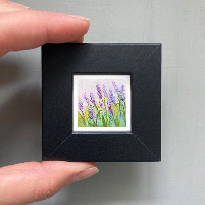 A hand is holding a small, square-shaped black frame containing a framed painting of purple flowers.